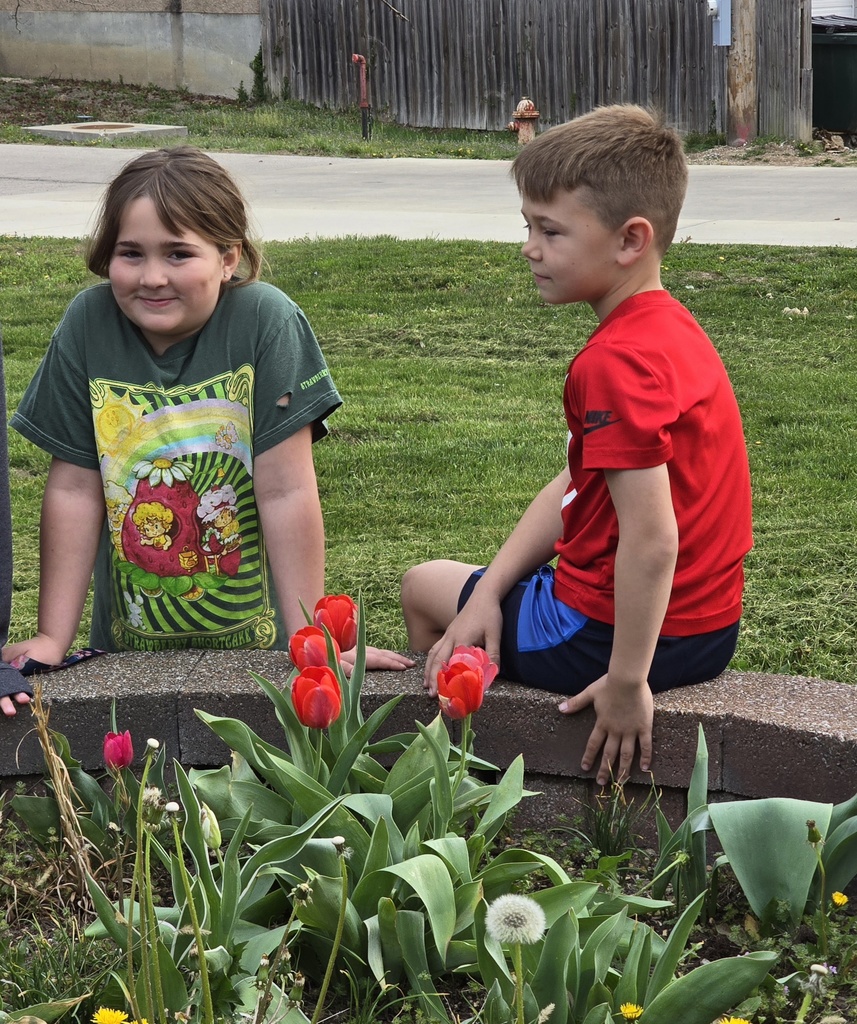 Two kids, one in a green shirt and the other in red, sit on a concrete edge. They face each other and smile in a garden with flowers and grass.