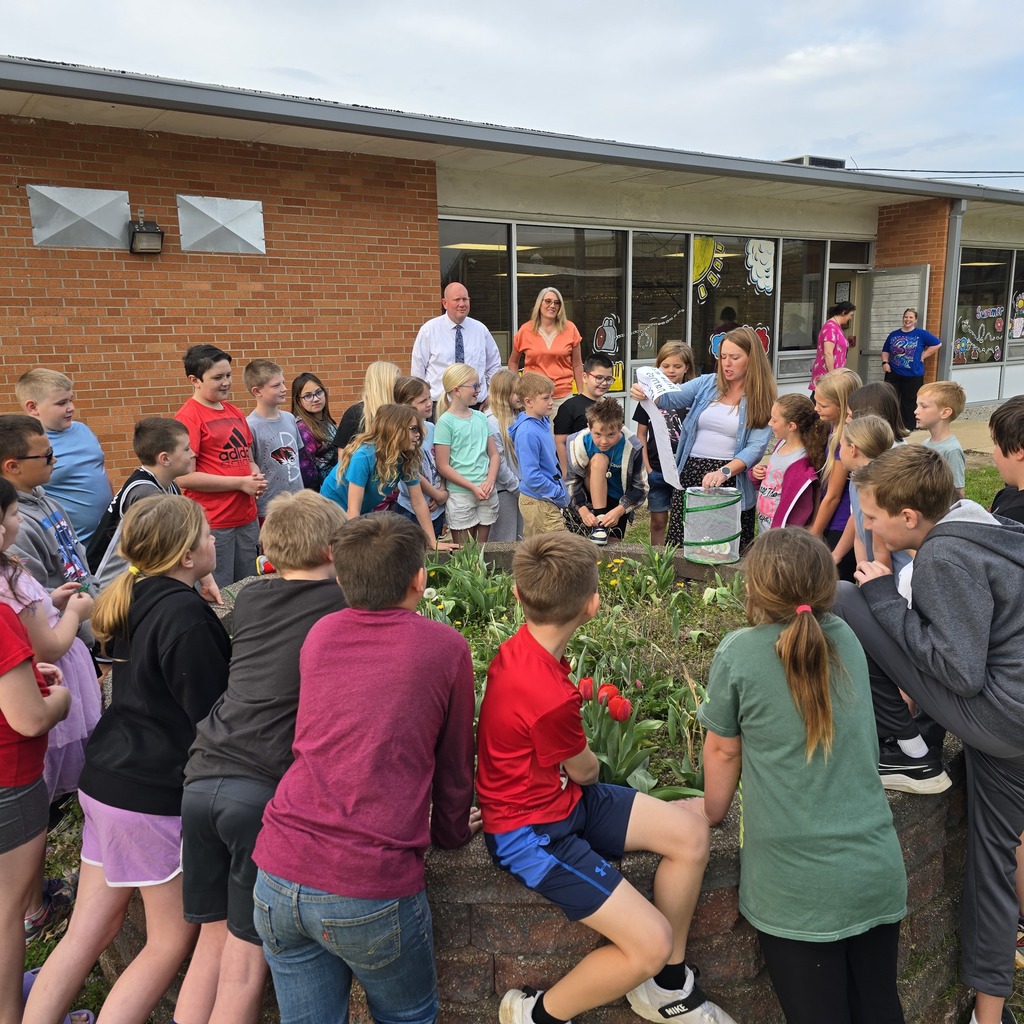 A group of children and adults gather around a flower bed in front of a brick building.
