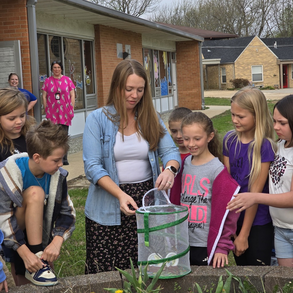 A woman and several children examine a bug trap while standing near a garden. A building with glass doors is behind them.