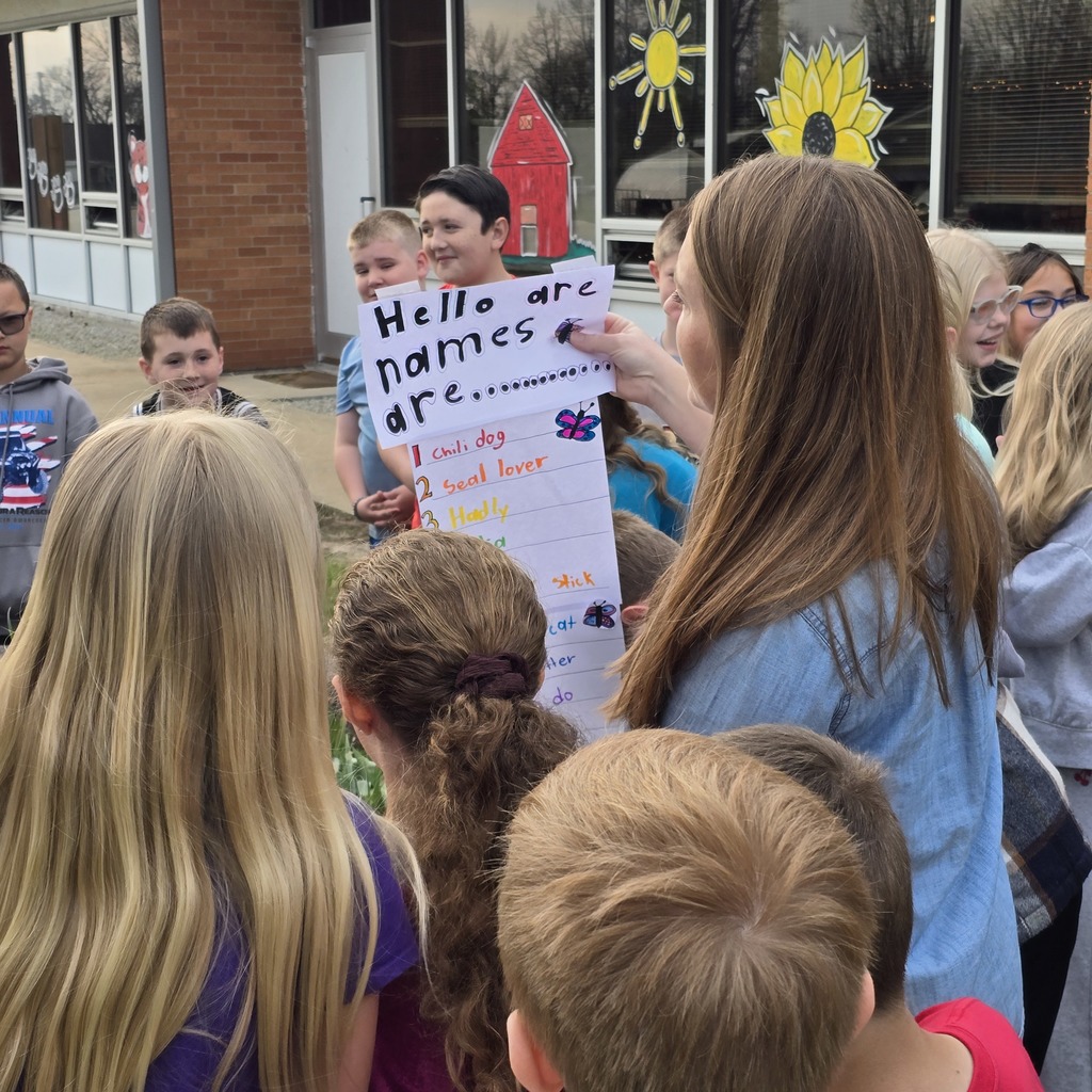 A group of children and adults outside a building, some holding signs with text. A woman holds a sign and speaks to a child.