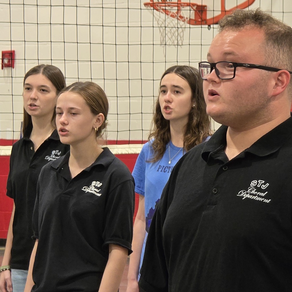Four individuals in black and blue shirts stand in a gymnasium, facing forward, with a basketball hoop in the background.