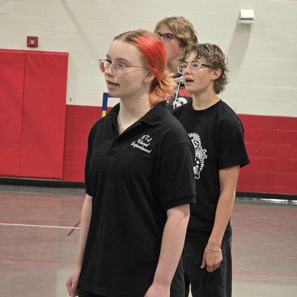 Three people in black shirts, one with red hair, stand in a gym with red and white walls.