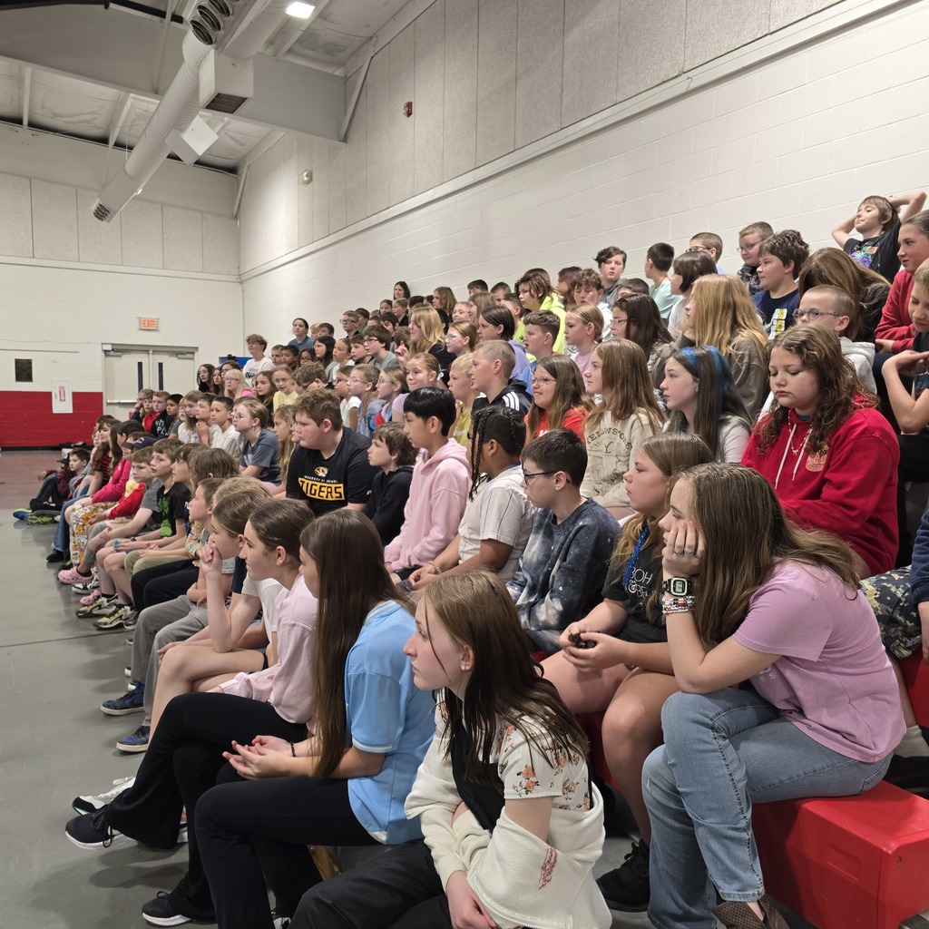 Many young people sit in red bleachers in a large room. They face the front, where a presentation is likely taking place.
