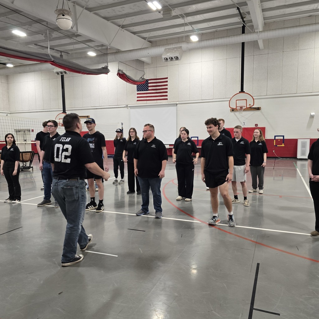A group of people in matching black shirts and sneakers are inside a gym, possibly practicing an activity.