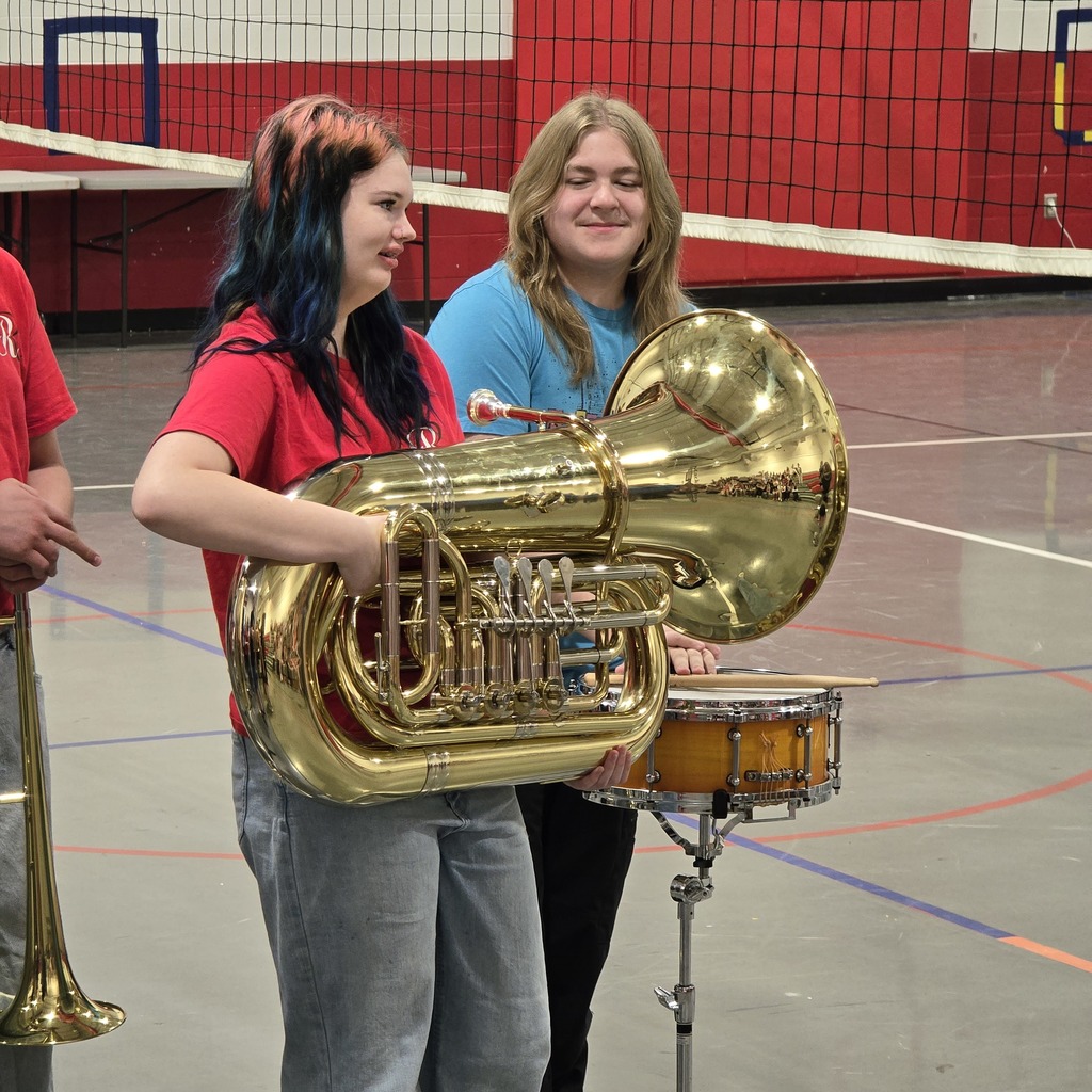 Two women in red shirts hold brass instruments in an indoor space with a red wall.