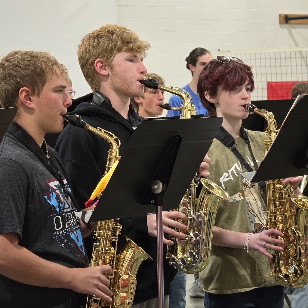 Four young musicians are playing saxophones, each holding one in both hands. They stand in front of music stands.