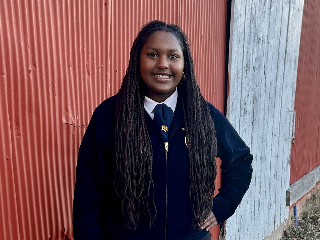 Person with long braided hair stands in front of red barn, smiling, wearing blue jacket and blue tie.