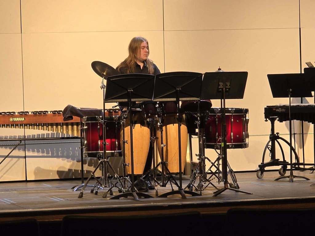 A person plays a drum set and other instruments on a stage with a plain backdrop.