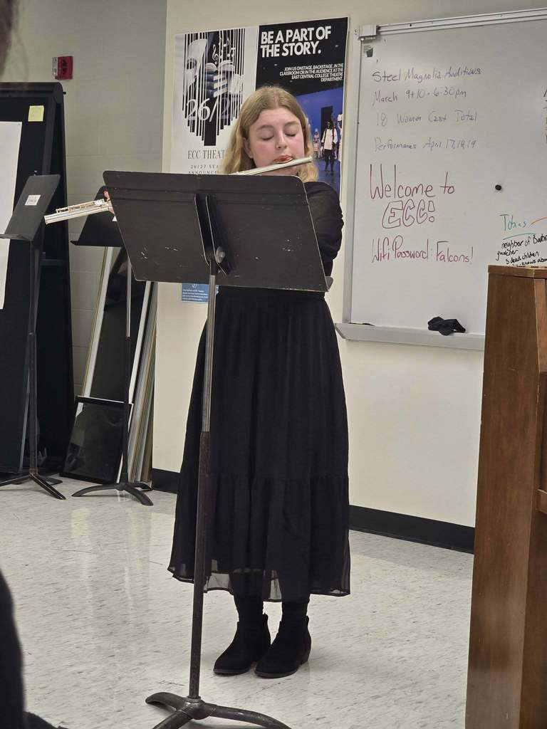A woman stands in a classroom, playing a flute on a music stand in front of a whiteboard.