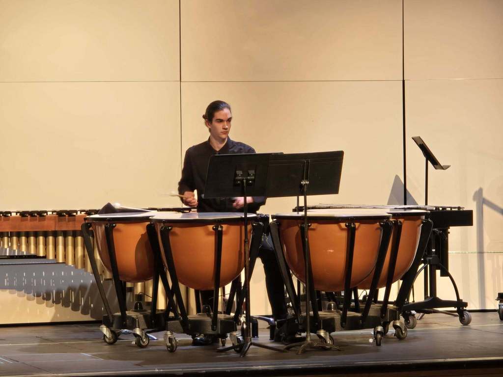 A person plays a set of timpani on a stage, with a music stand in front and other percussion instruments in the background.