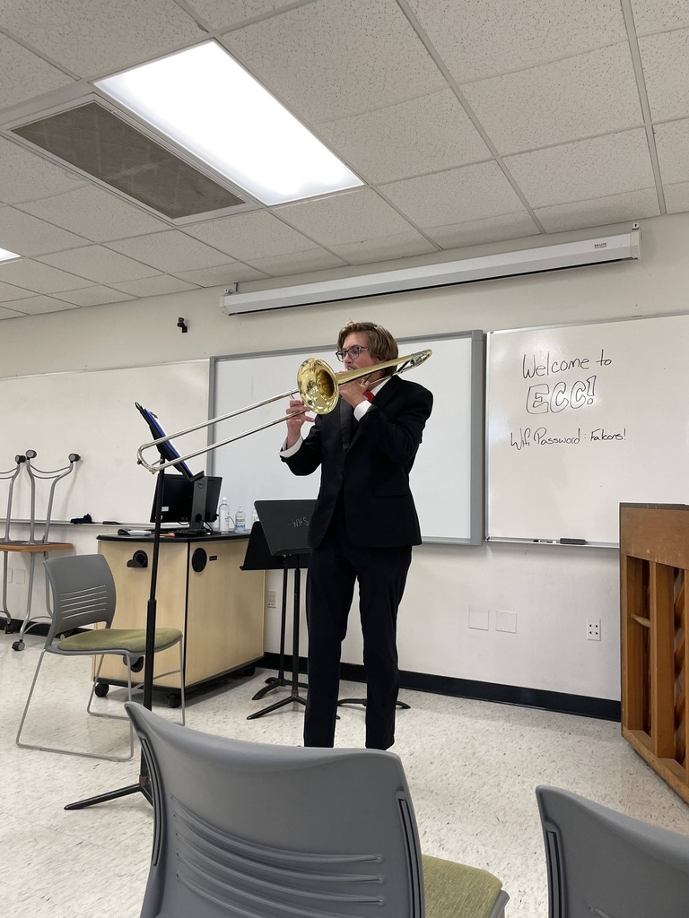 A man stands in a classroom playing a trombone in front of a whiteboard and chairs.