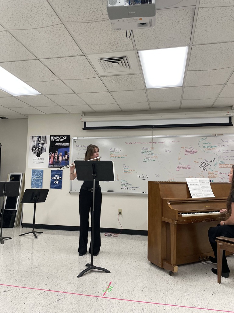 A person plays a flute while standing next to a music stand in a classroom. Another person plays piano.