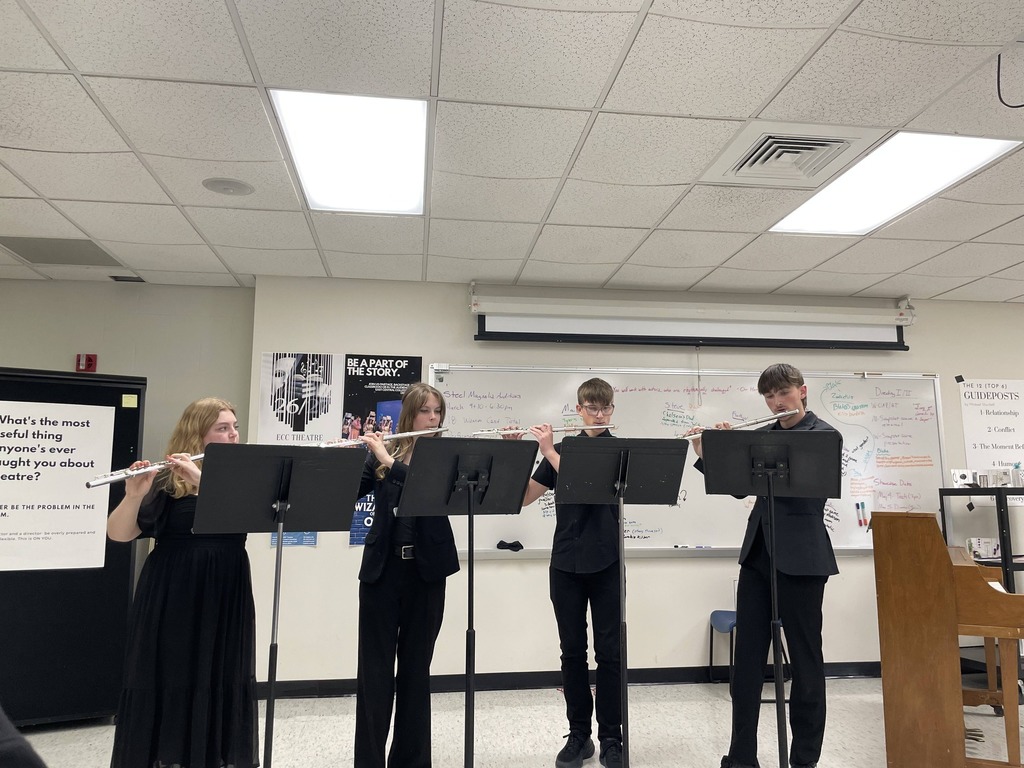 Four people stand in a row, playing flutes in a room with a piano, whiteboards, and ceiling lights.
