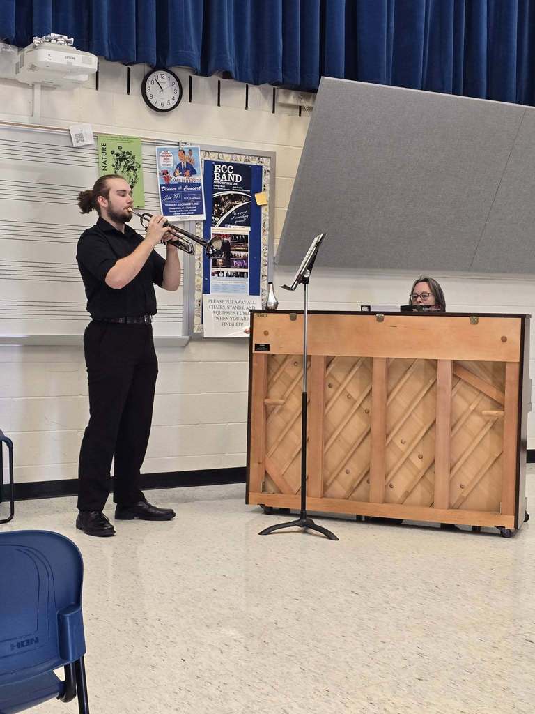 A person in black plays a trumpet in a classroom. A piano is behind them, with a woman seated in front.