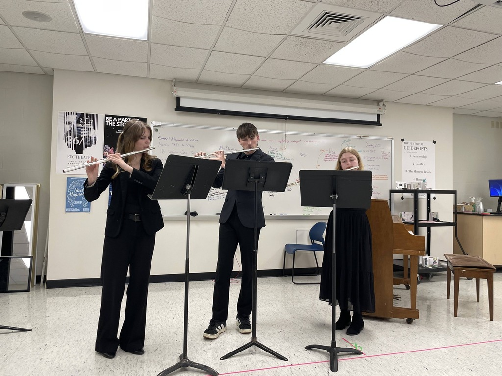 Three individuals in formal attire, one playing the flute and the other two playing clarinets, in a room with a piano and whiteboard.