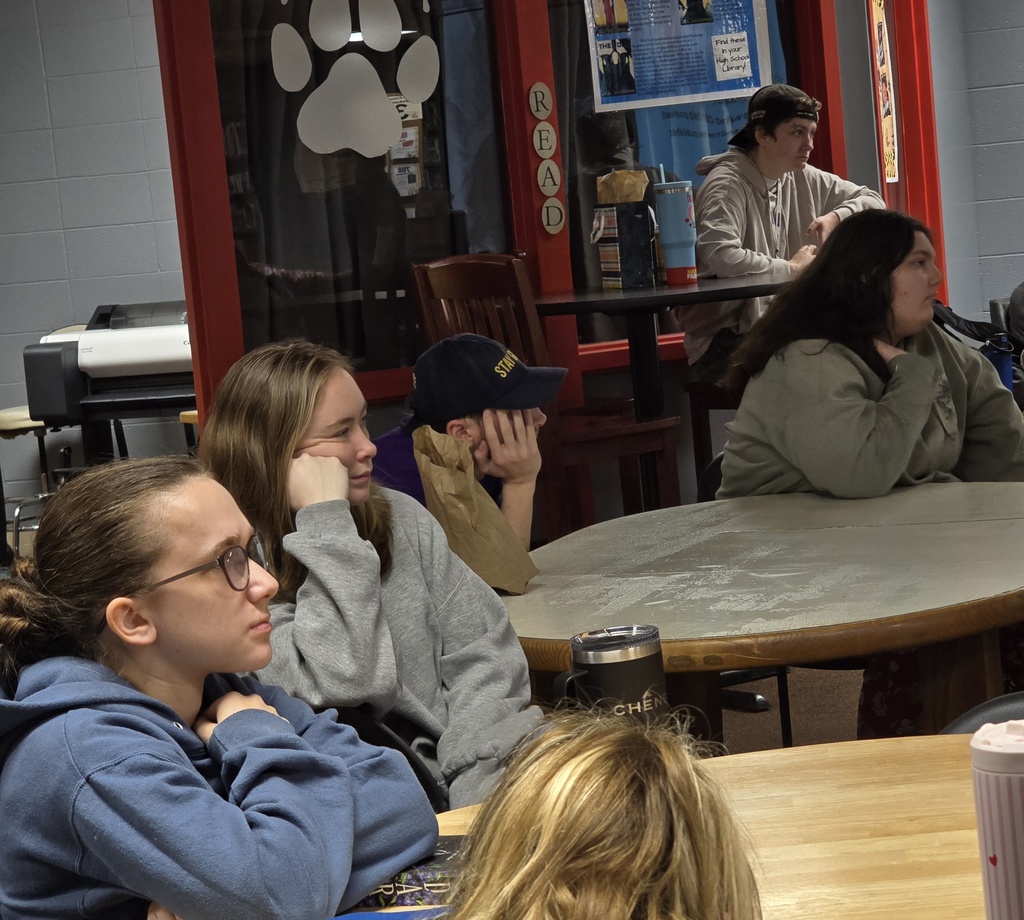 Students seated around tables inside a room with a paw print on the wall. Some students wear glasses.