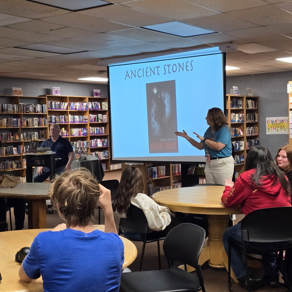 Teacher stands in front of presentation board with students at tables.