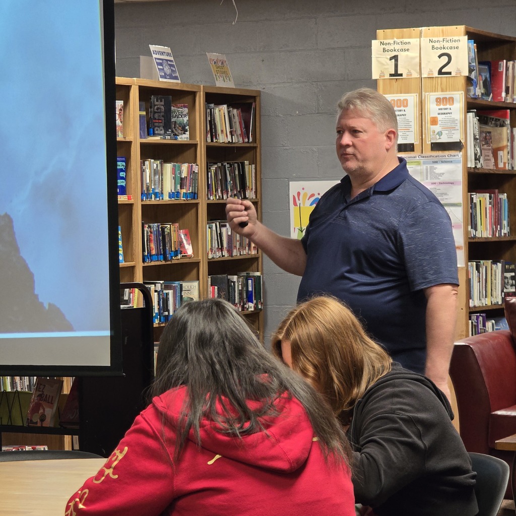 Man standing in front of a screen, showing a presentation. Two women in front, one in red and one in black.
