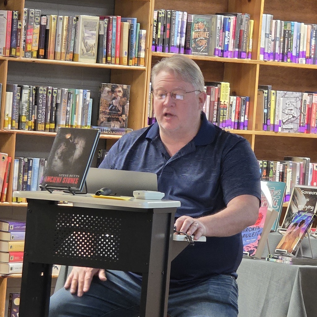 Man wearing glasses and a dark blue shirt giving a presentation in front of a table in a bookstore.