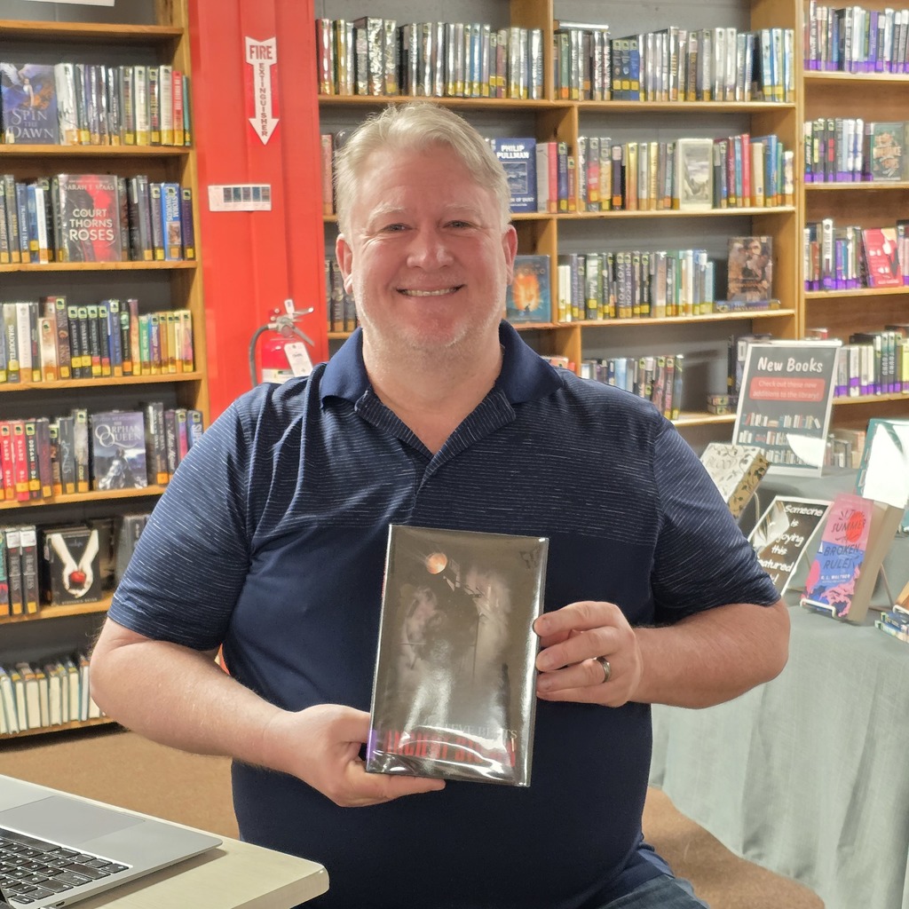 A man holds a book in a library. He smiles, and shelves filled with books are behind him.