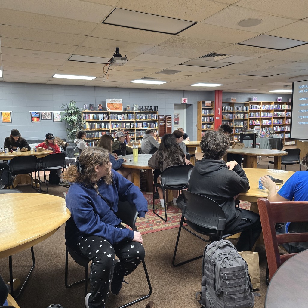 A room with tables and chairs has people seated. The tables have books and items on them. The floor is carpeted.