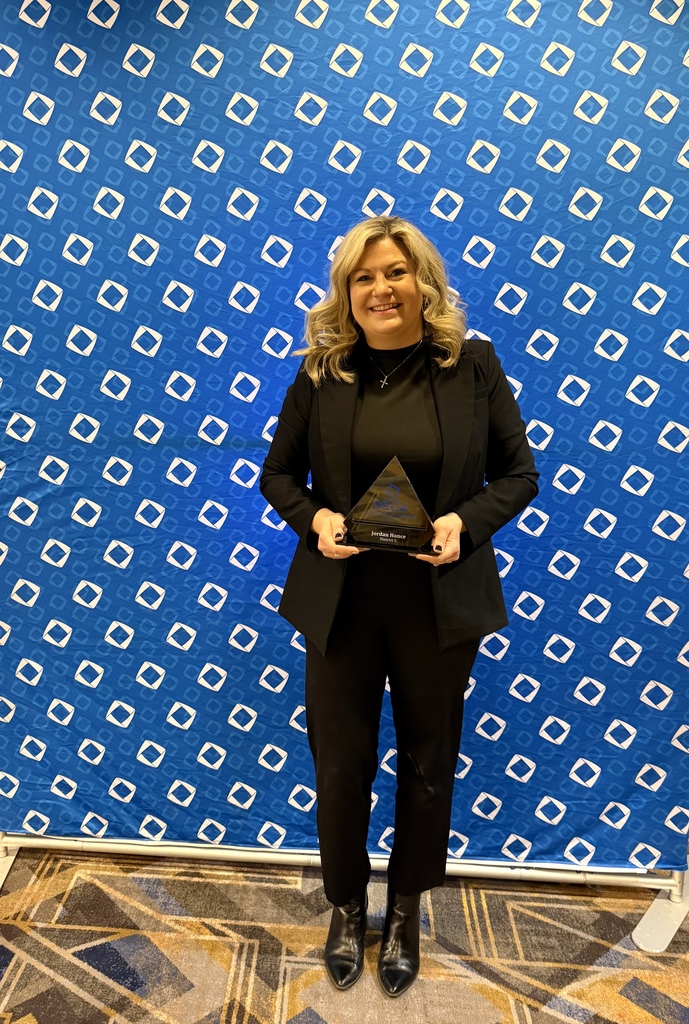 A woman in a black suit holds a trophy in front of a blue diamond-patterned wall.