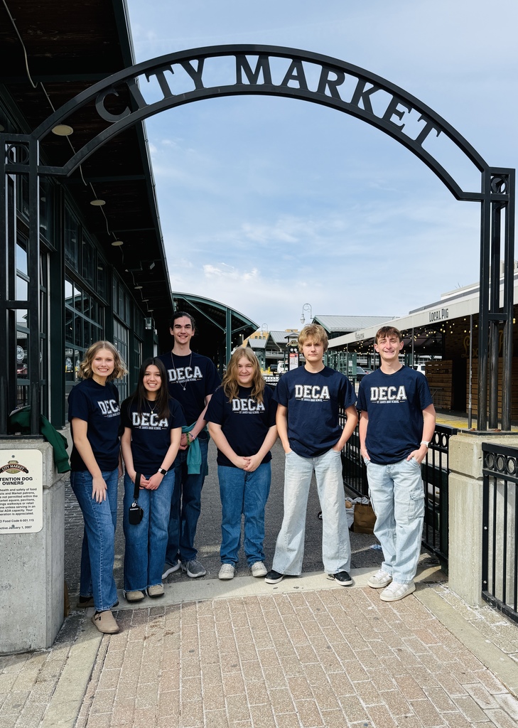 Seven individuals stand together under a sign that reads "CITY MARKET" in white letters. They wear matching blue shirts.