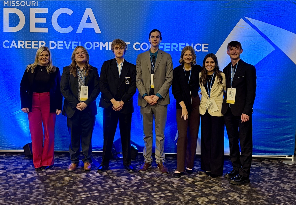 Seven people stand in front of a blue backdrop with the words "DECA" and "Career Development Conference".