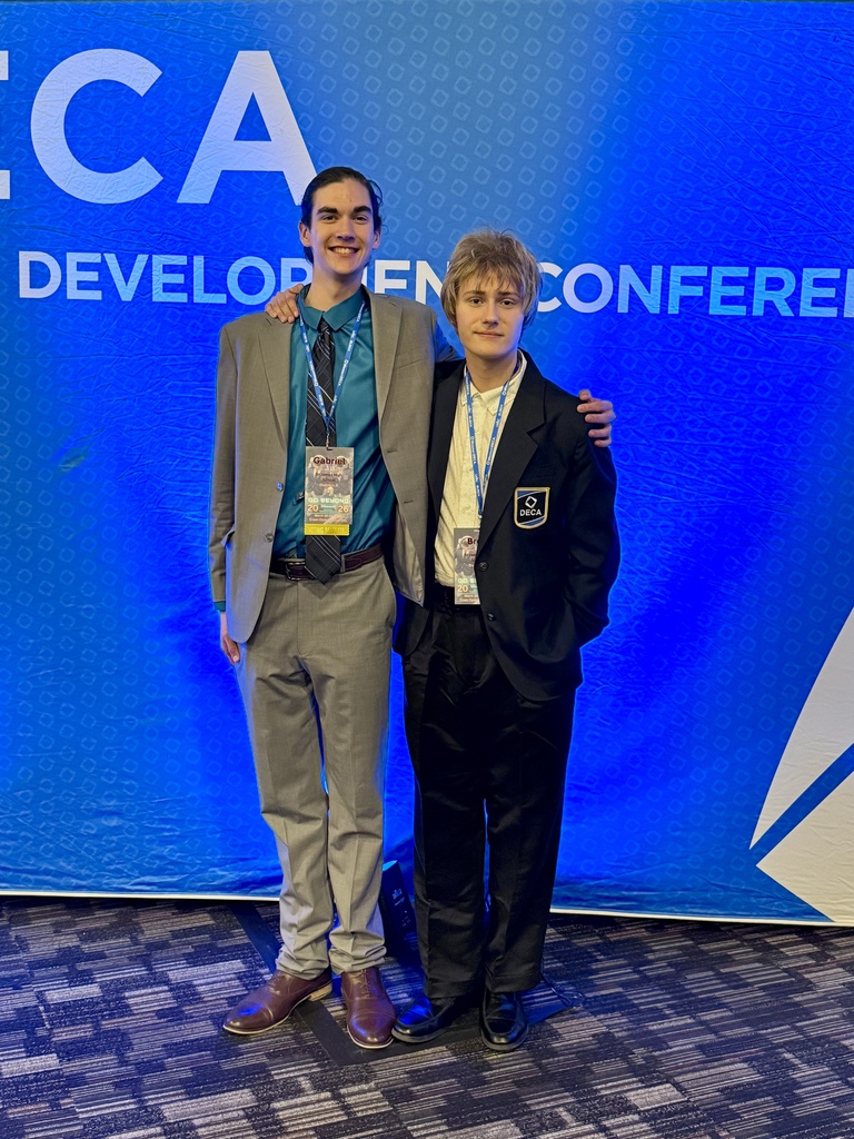 Two individuals in formal wear pose in front of a blue backdrop displaying white text.
