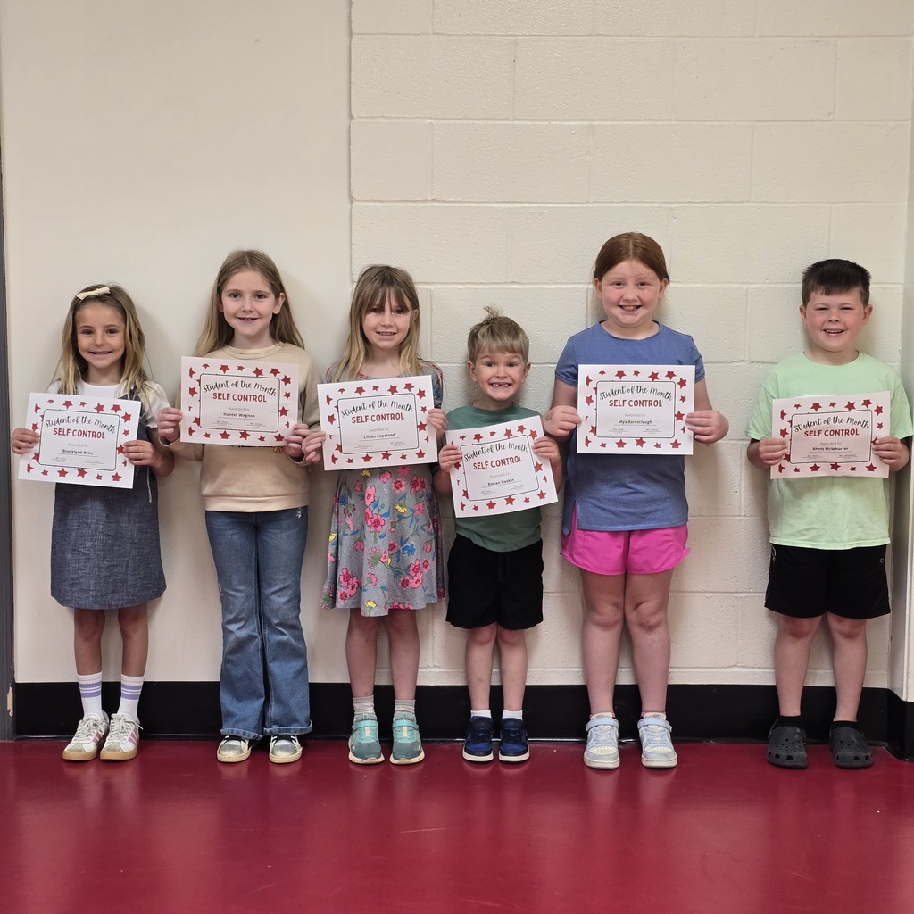 Seven children stand in a row, holding certificates with red patterns. They are in a room with red flooring.