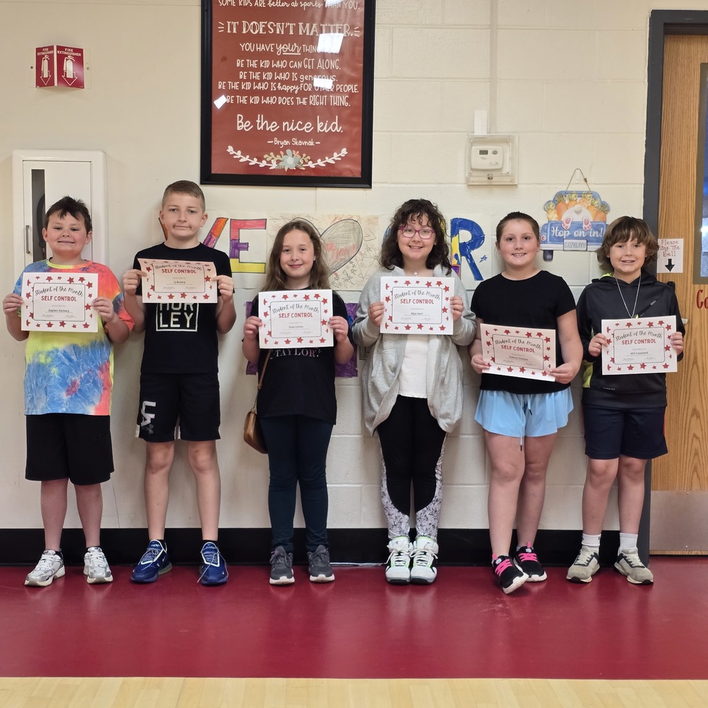 Six children hold certificates in a classroom. Behind them is a framed bulletin board with writing and a red border.