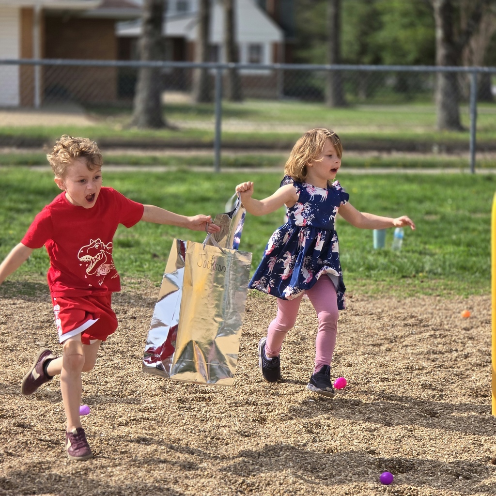 Two children run in an outdoor park, holding bags, with a grassy area, fence, and building in the background.