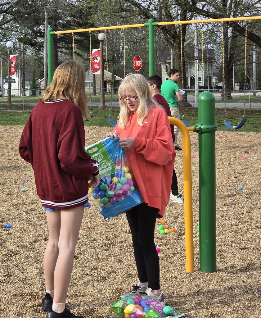 Two young women in a playground. One holds a bag of Easter eggs. Several eggs are scattered around.