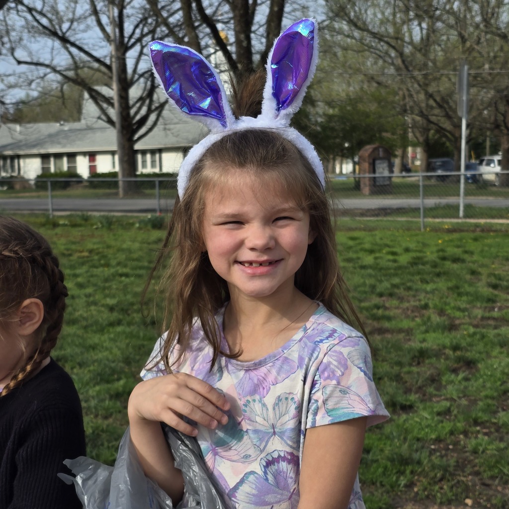A girl with bunny ears smiling and standing in a park with a fence and trees.