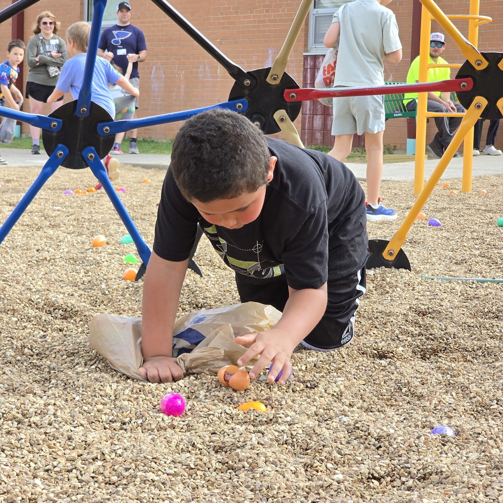 A boy bends over, picking up small orange balls from the ground at a playground. Other children and adults are in the background.