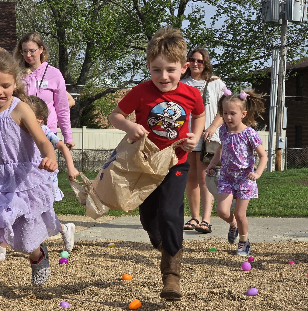 Children in dresses and shirts run on a gravel area, one carrying a brown bag. Trees and a fence in the background.