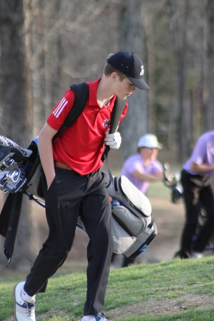 A golfer in a red shirt and black cap walks with a golf bag on a grassy slope, trees in the background.