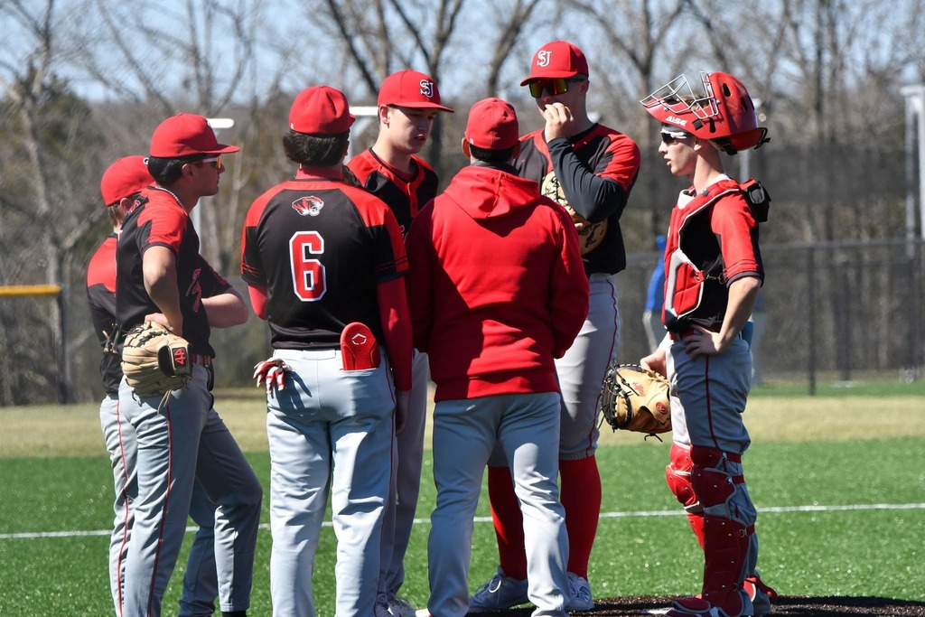 Baseball team in red and black uniforms, hats, and equipment gather for a discussion on a field with green grass and trees.