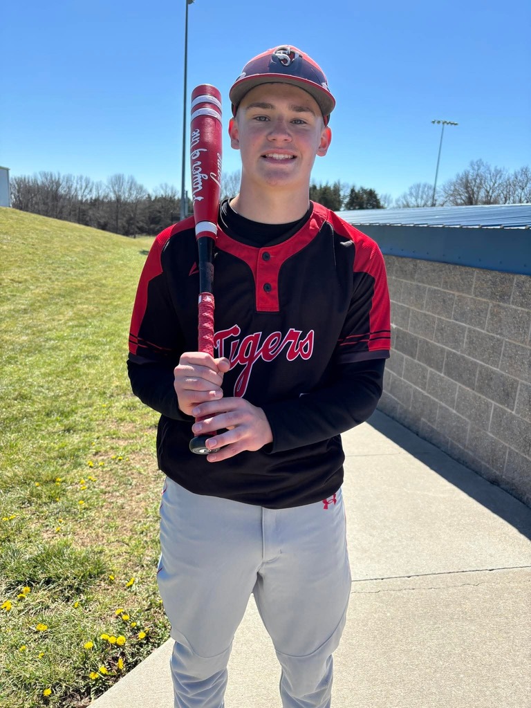 A young man in a baseball uniform holds a bat, smiling. Behind him is a wall and a grassy area.