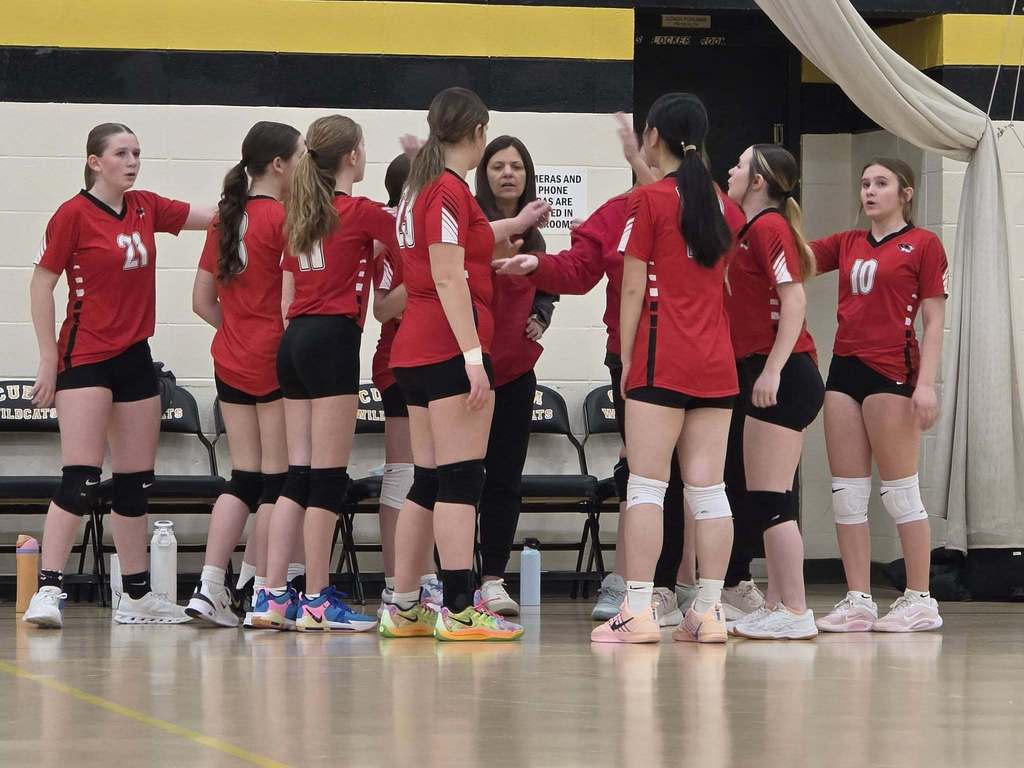 A group of female volleyball players in red uniforms, gathered for a huddle with coaches on a court.
