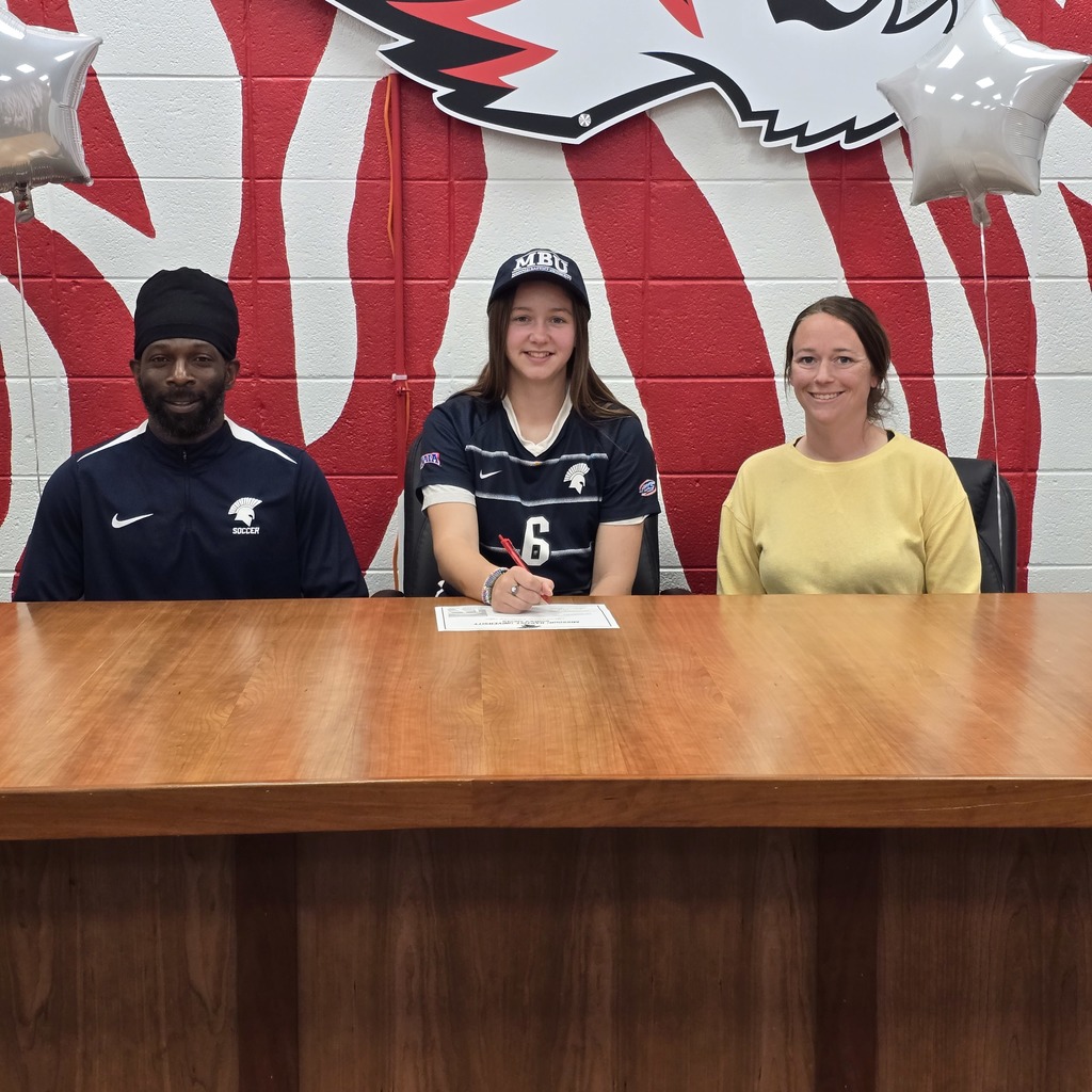 Three individuals seated at a table, one holding a pen, with a red and white backdrop displaying a school emblem.