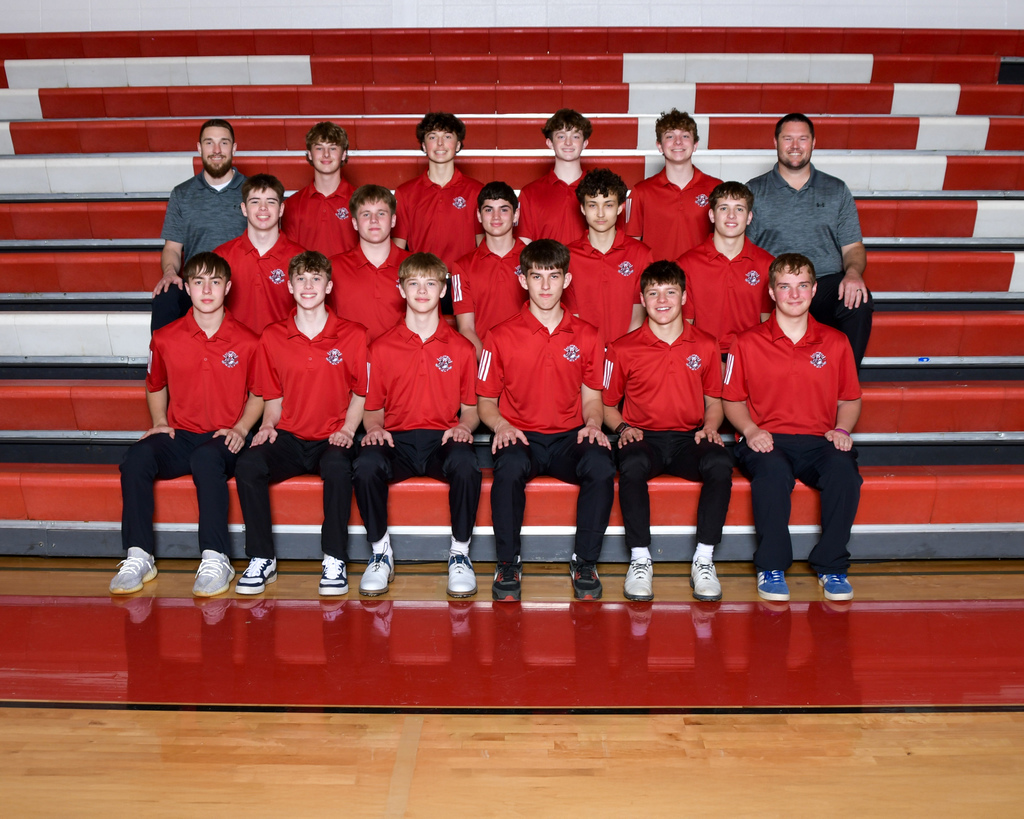 A group of teenage boys in red shirts and black pants pose on red bleachers, with two men in gray shirts.