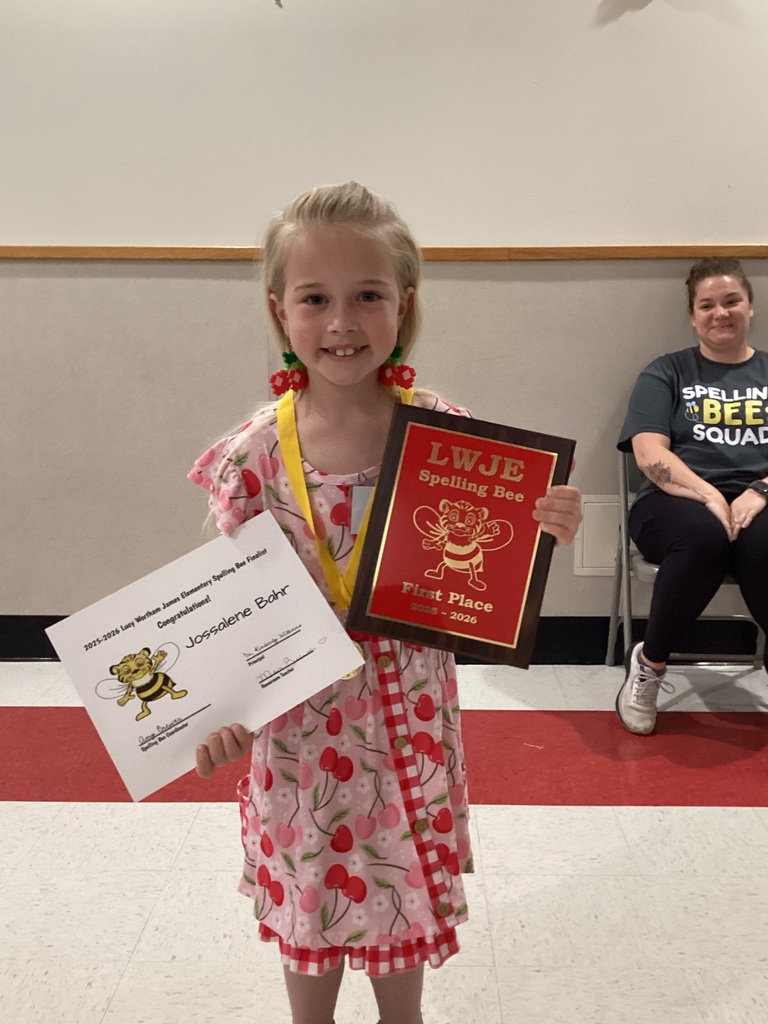 A young girl in a pink dress holds a certificate and a red plaque, smiling at the camera. Behind her, a woman sits on a chair, wearing a black T-shirt.