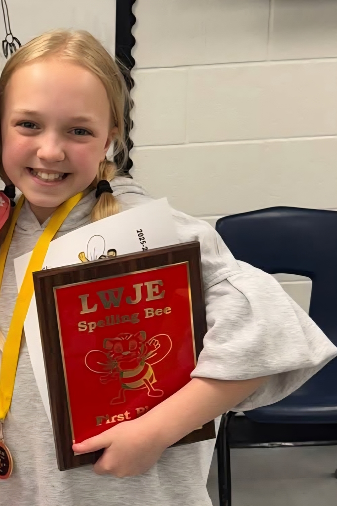 A girl holds a framed award for first place in a spelling bee. She smiles, with her gray sweatshirt and yellow medal visible.