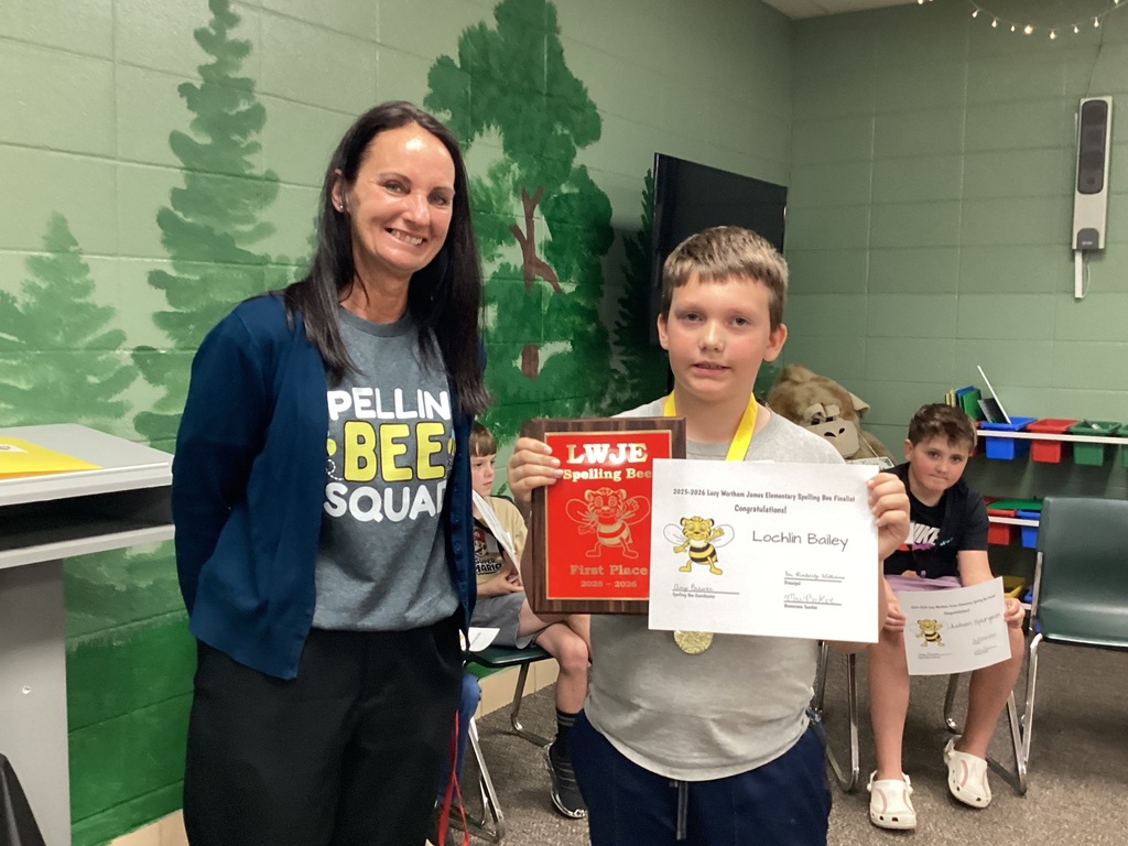Woman and boy standing, smiling, and holding certificates. The boy wears a medal. Green wall with tree images in the background.