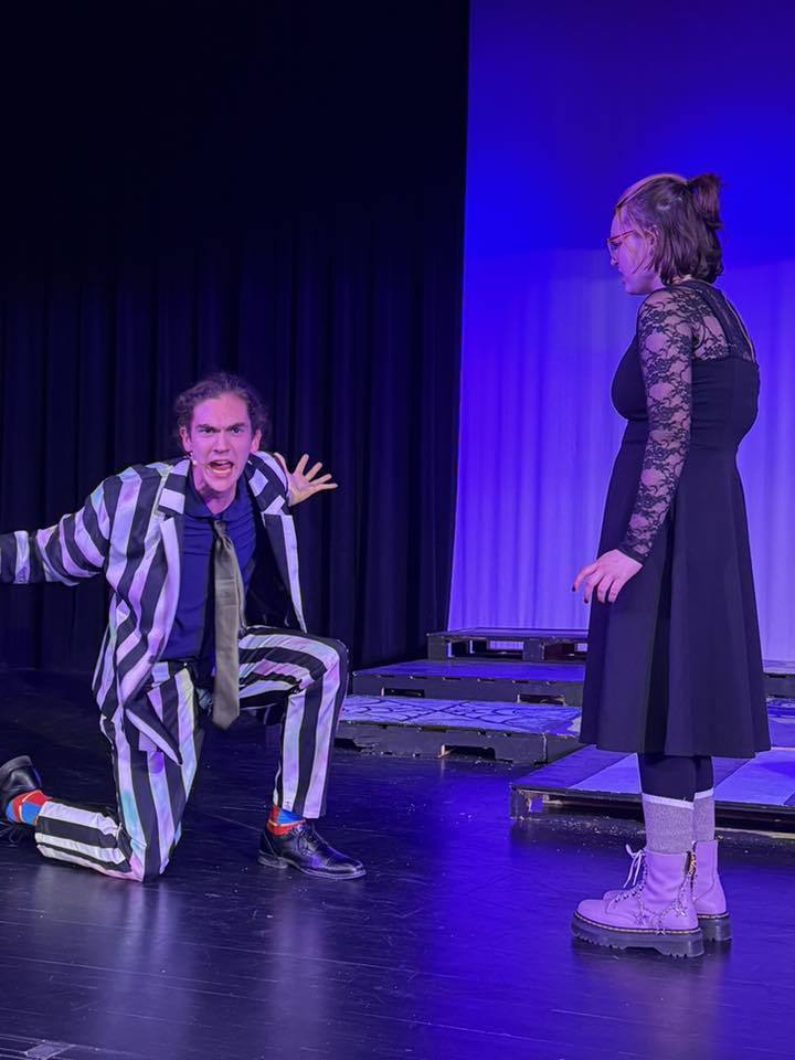 Person in striped suit kneels on a stage; another in a long dress stands nearby. Dark background with blue lighting.