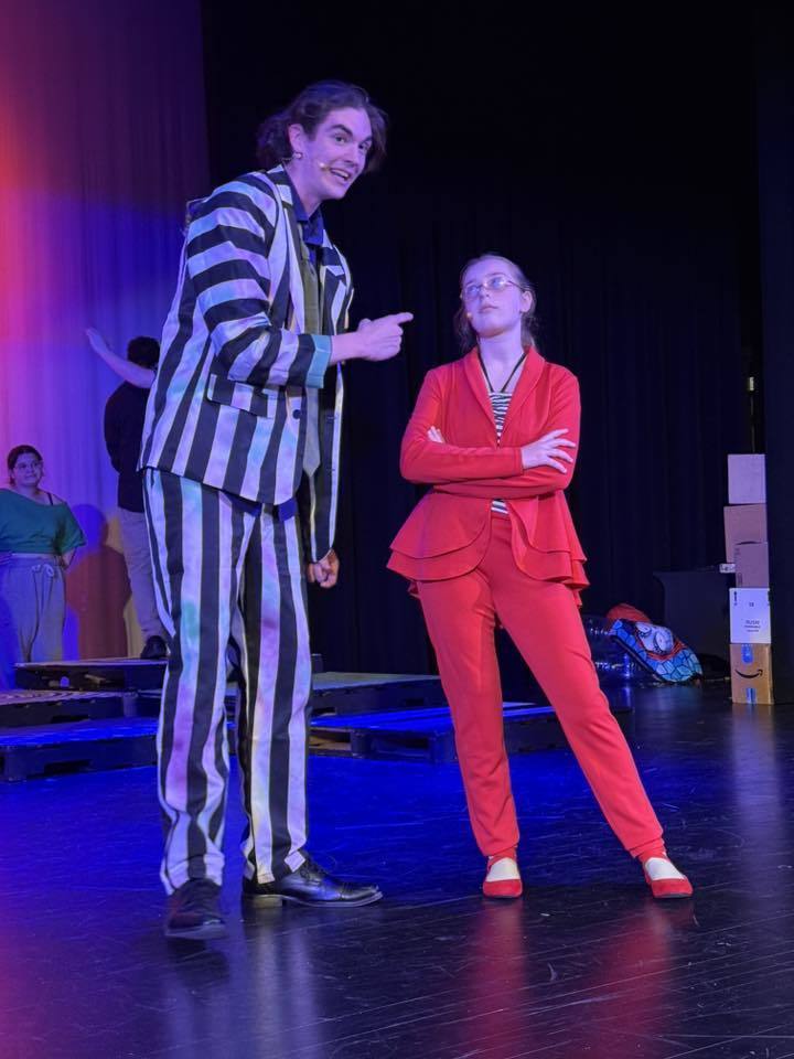 Two people in a performance. One wears a striped suit, the other a red outfit. They stand on a stage.
