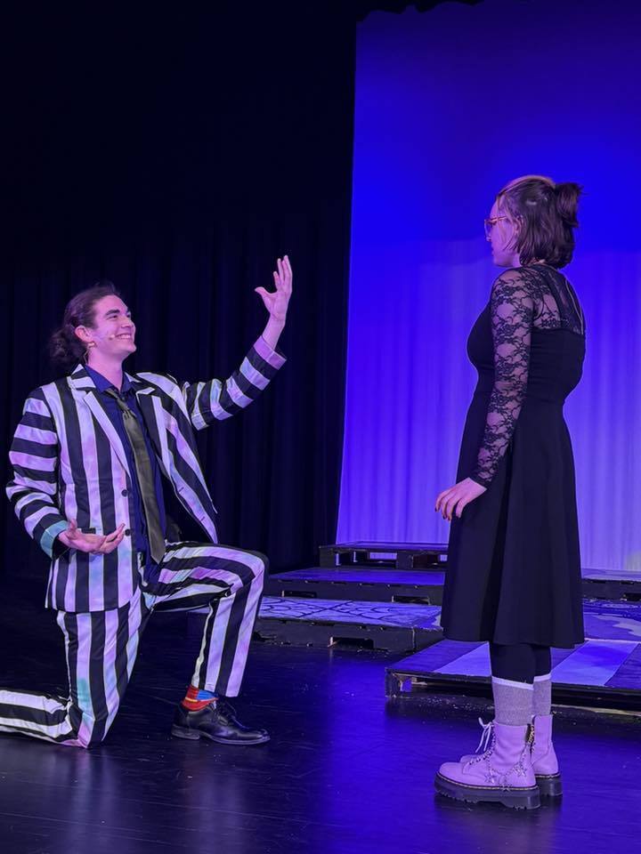 A person kneeling in a striped suit gestures to another person in a black dress on a stage.