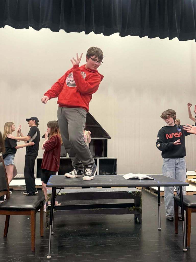 People practice on a stage. A man jumps above a table. Other people surround him, some clapping.