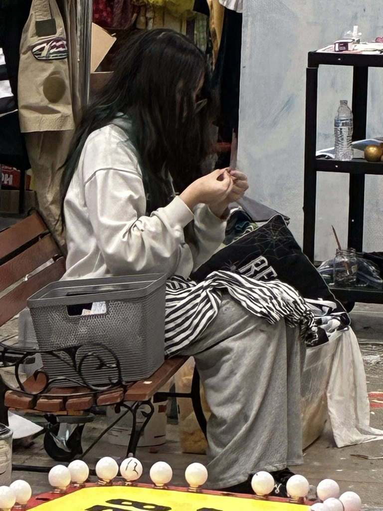 Person seated on a bench with a bag, working on a craft project with various objects around.